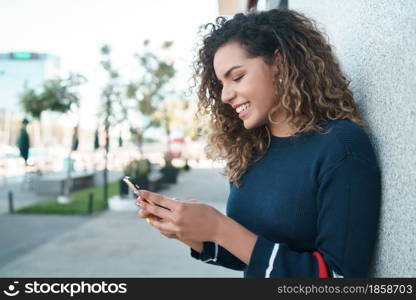 Young latin woman smiling while using her mobile phone outdoors on the street. Urban concept.