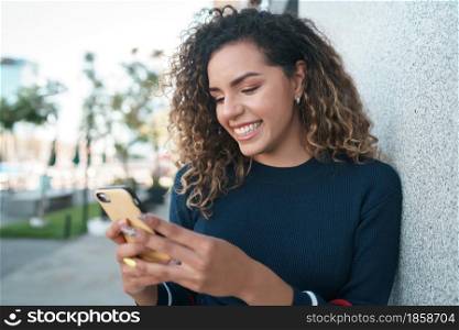 Young latin woman smiling while using her mobile phone outdoors on the street. Urban concept.