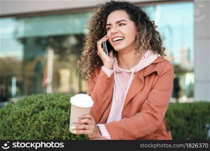 Young latin woman talking on the phone while sitting on a bench outdoors in the street. Urban concept.