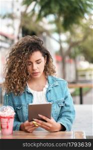 Young latin woman using a digital tablet while drinking a cold drink beverage at a coffee shop outdoors on the street.