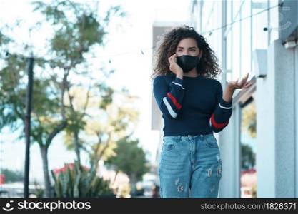 Young latin woman using a face mask while talking on the phone outdoors in the street. New normal lifestyle. Urban concept.