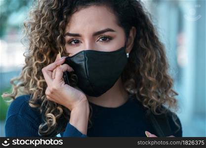 Young latin woman using a face mask while talking on the phone outdoors in the street. New normal lifestyle. Urban concept.