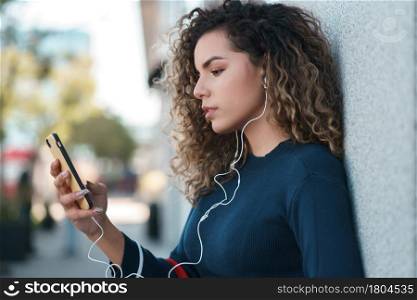 Young latin woman using her mobile phone while standing outdoors at the street. Urban concept.