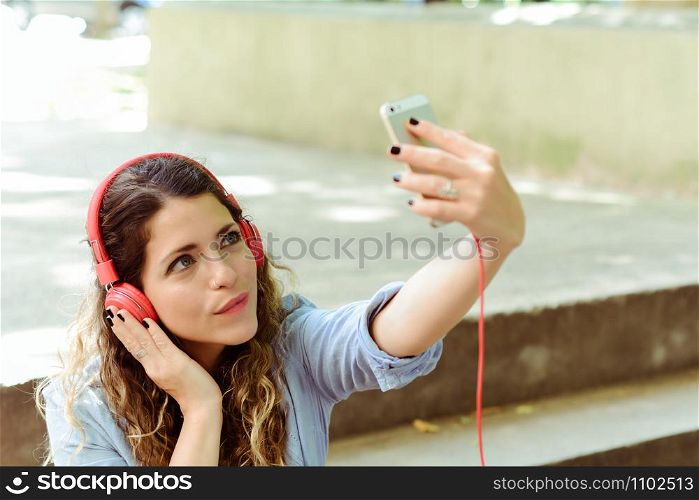 Young latin woman with red headpones making a selfie with mobile phone. Outdoors