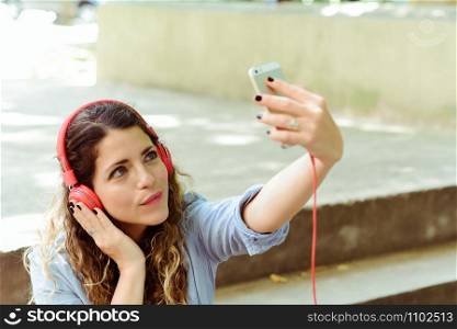 Young latin woman with red headpones making a selfie with mobile phone. Outdoors
