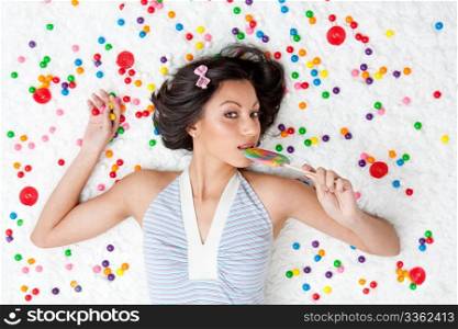 Young Latina woman laying on ruffled cloud like floor between colorful bubblegum balls eating a lollipop