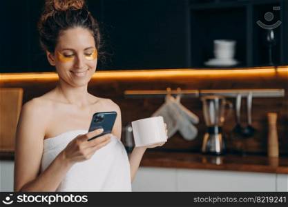 Young latina woman reading messages on smartphone. Happy girl wrapped in towel with under eye patches chatting in social networks, shopping online after morning shower on kitchen at home.. Girl with patches under eyes chatting in social networks, shopping online by phone in the morning