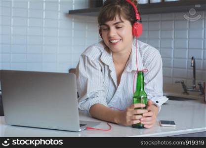 Young latn woman with beer and using her laptop pc in the kitchen at home.