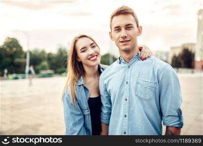 Young laughing couple hugs in summer city park. Smiling teenagers poses together, cityscape on background