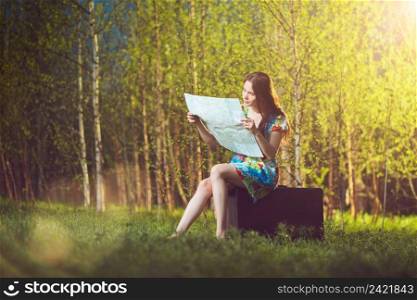 Young lost woman sitting on a suitcase and reading a map in woods  lens flare