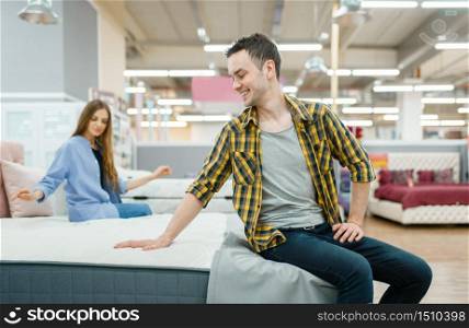 Young love couple checks mattress in furniture store showroom. Man and woman looking samples for bedroom in shop, husband and wife buys goods for modern home interior. Couple checks mattress in furniture store showroom