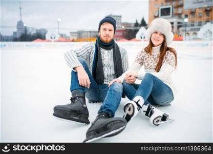 Young love couple in skates sitting on ice, skating rink. Winter ice-skating on open air, active leisure. Love couple in skates sitting on ice, skating rink