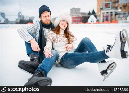 Young love couple in skates sitting on ice, skating rink. Winter ice-skating on open air, active leisure. Love couple in skates sitting on ice, skating rink