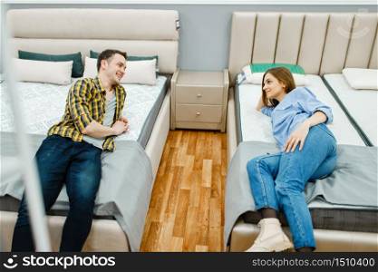 Young love couple lying in bed in furniture store showroom. Man and woman looking samples for bedroom in shop, husband and wife buys goods for modern home interior. Love couple lying in bed, furniture store showroom