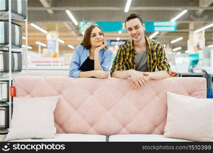 Young love couple poses at the pink couch in furniture store. Man and woman looking samples for bedroom in shop, husband and wife buys goods for modern home interior. Couple poses at the pink couch in furniture store