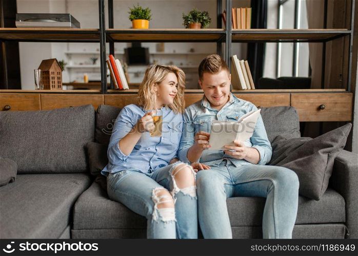 Young love couple sitting on couch at home. Husband and wife relax in living room. Happy family together, man reading a book, wife drinks tea