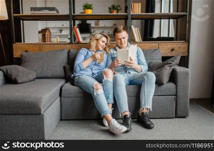 Young love couple sitting on couch at home. Husband and wife relax in living room. Happy family together, man reading a book, wife drinks tea. Young love couple sitting on couch at home