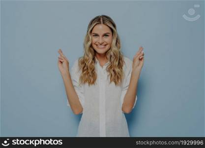 Young lovely caucasian blonde female with beaming smile raising hands and crossing fingers with hope and luck gesture, anticipating important news, isolated over blue studio background. Young lovely blonde female crossing fingers with luck gesture