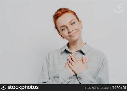 Young lovely red haired woman wearing shirt holding folded hands on chest and feeling grateful and thankful and support, looking at camera with charming smile, isolated on studio grey background. Young lovely red haired woman holding folded hands on chest and feeling grateful and thankful