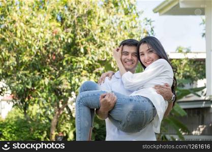 Young loving couple have fun in park. Caucasian boyfriend carry on his asian girlfriend in arms in front yard of house.