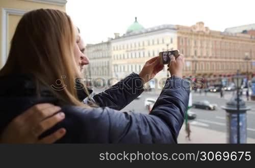 Young loving couple outdoor using smartphone to take funny pictures of themselves. City view in background