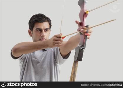 Young male archer aiming bow and arrow against gray background