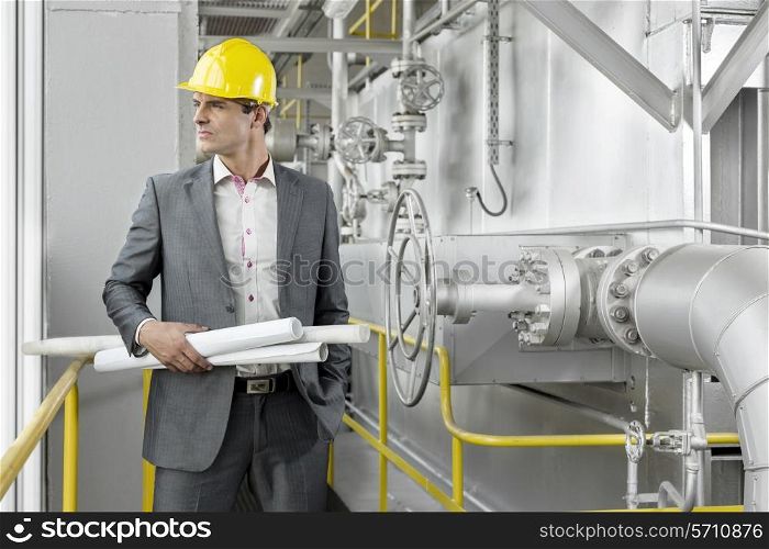 Young male architect holding rolled up blueprints by industrial machinery