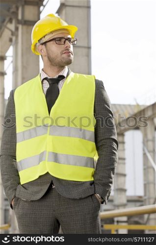 Young male architect in protective workwear standing hands in pockets at construction site