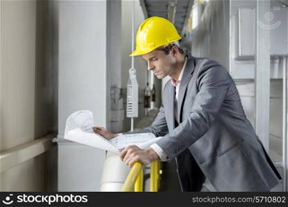 Young male architect reading blueprint while leaning on railing in industry