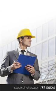 Young male architect with clipboard looking away in front of office building