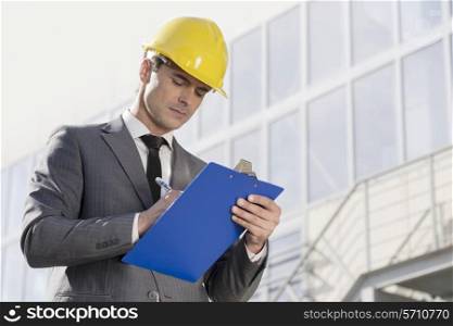 Young male architect writing on clipboard against office building