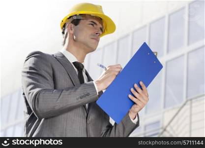 Young male architect writing on clipboard while looking away outside building