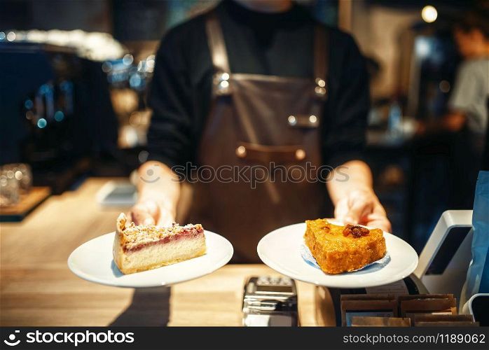 Young male barista holding in hands plates with sweet dessert, cafe counter on background. Professional coffee preparation by barman in cafeteria, bartender occupation. Barista holding in hands plates with sweet dessert
