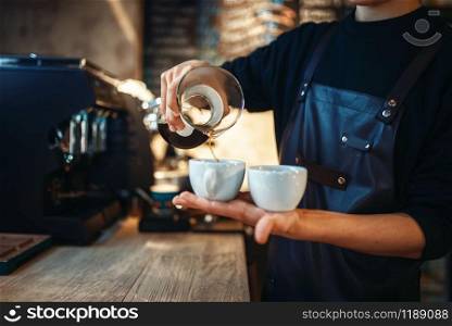 Young male barista in black apron holds in hands two cups of fresh coffee, cafe counter on background. Professional espresso preparation by barman in cafeteria, bartender occupation. Young male barista holds two cups of fresh coffee