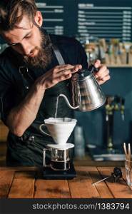 Young male barista pouring boiling water from kettle to drip coffee maker on wooden table. Barista wearing dark uniform. Tools and equipment for making Drip Brew coffee on wooden table.