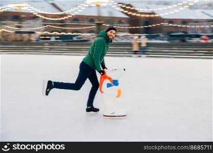 Young male begginer learns skating with help of special figure, leans at it, being on ice rink covered with snow, has cheerful expression, poses in camera. Man uses special equipment for skating