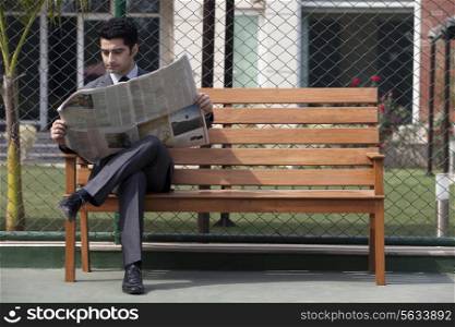 Young male business executive reading newspaper while sitting on bench in tennis court