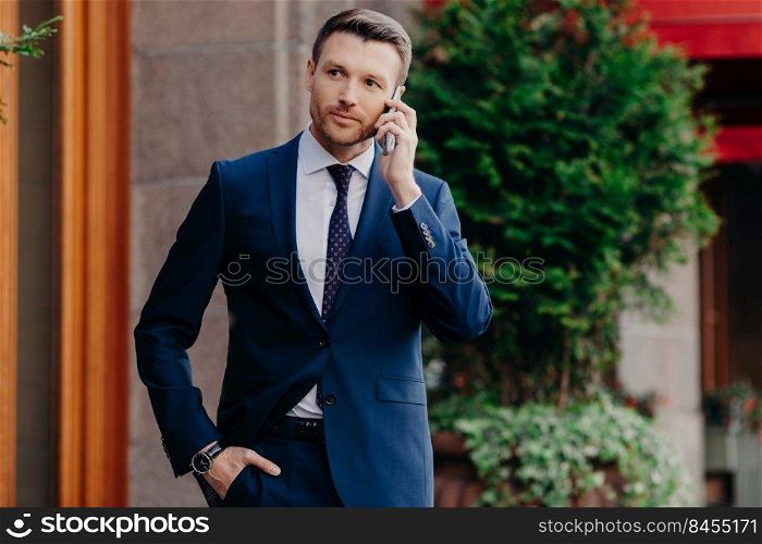 Young male CEO has telephone conversation, looks confidently into distance, dressed in elegant suit, poses outdoor, uses modern cell phone. Successful businessman has talk with business partner