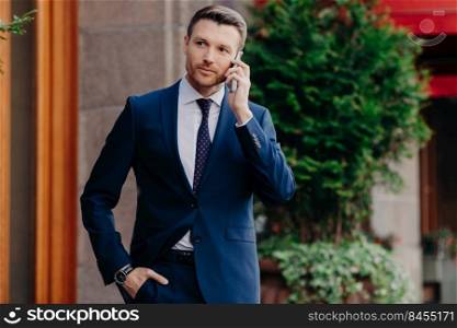 Young male CEO has telephone conversation, looks confidently into distance, dressed in elegant suit, poses outdoor, uses modern cell phone. Successful businessman has talk with business partner