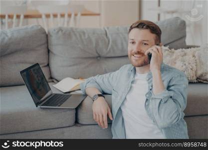 Young male company employee working remotely from home, calling his boss to tell him good news about projects progress, resting hand on couch with laptop and data while sitting on carpet. Young male company employee working remotely from home calling boss to tell good news