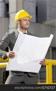 Young male engineer holding blueprint while looking away at construction site