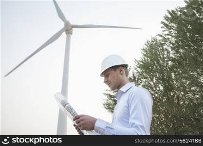 Young male engineer in a hardhat looking down at a blueprint in front of a wind turbine