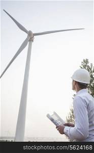 Young male engineer in a hardhat looking up at a wind turbine, blueprint in hand