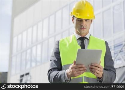 Young male engineer in reflector-vest and hardhat using digital tablet outside industry