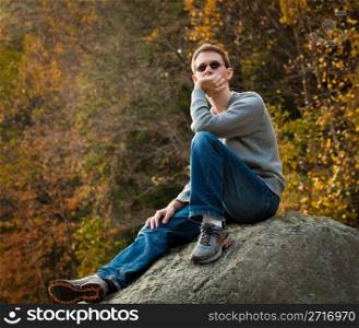 Young male hiker in jeans pondering while sitting on a boulder in front of fall leaves