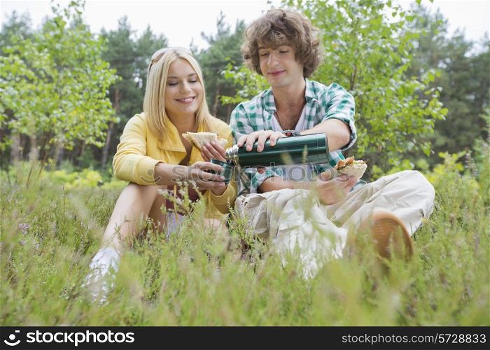 Young male hiker pouring coffee for woman while relaxing in field