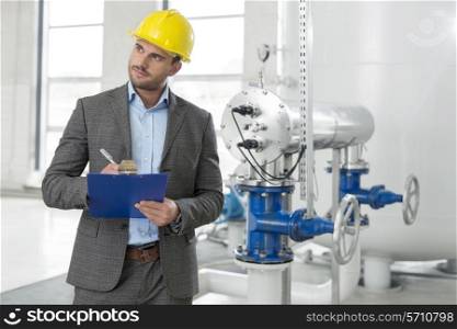 Young male inspector writing on clipboard while looking away in industry