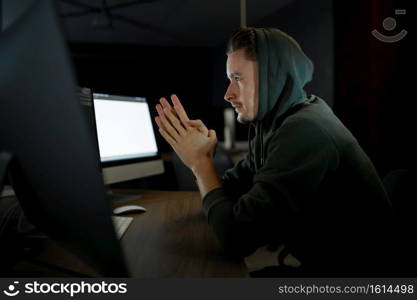 Young male internet hacker in hood sitting at monitors. Illegal web programmer at workplace, criminal occupation. Data hacking, cyber security. Young internet hacker in hood sitting at monitors