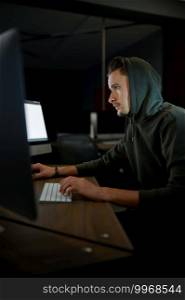 Young male internet hacker in hood sitting at monitors. Illegal web programmer at workplace, criminal occupation. Data hacking, cyber security. Young internet hacker in hood sitting at monitors