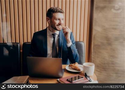 Young male investor dressed in blue suit looking outside window while sitting in cafe hearing good news about investments from his partners via online call on laptop, coffee and croissant on his table. Male investor in blue suit talking with business partner online through laptop in restaurant